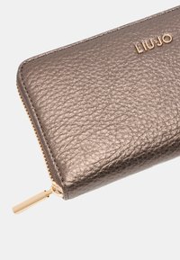 Portafoglio in pelle metallizzata bronzo con finitura testurizzata, dotato di zip dorata e logo "LIU JO" in lettere dorate su uno sfondo chiaro.