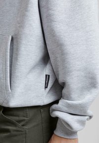 Grå genser laget av mykt stoff, med en frontlomme og et synlig svart merke med teksten "PROHIBITED" nær kanten. Ribbestrikkede ermer.