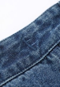 Tela de mezclilla en un azul profundo, con un logo cosido distintivo y una superficie texturizada. La cinturilla presenta un corte limpio y una costura uniforme.