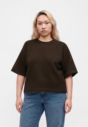 Ung kvinde med langt blond hår iført en mørkebrun kortærmet top og blå jeans, står foran en ensfarvet hvid baggrund.
