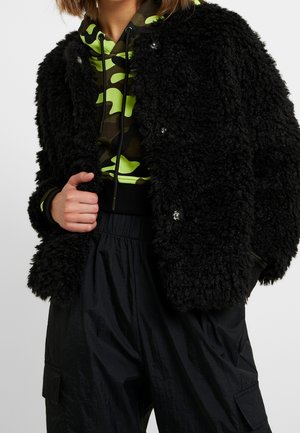 Persona con chaqueta negra texturizada sobre sudadera con capucha de camuflaje verde y negro, con pantalones cargo negros, mano sujetando el borde de la chaqueta.
