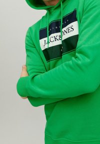 Muž se zkříženýma rukama, oblečený v jasně zelené mikině s kapucí, na které je černobílé a zelené logo "JACK & JONES" na hrudi, na jednobarevném pozadí.