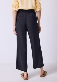 Black wide-leg pants with an elastic waistband, featuring two back pockets and a smooth, lightweight fabric. Model wearing brown slip-on shoes.
