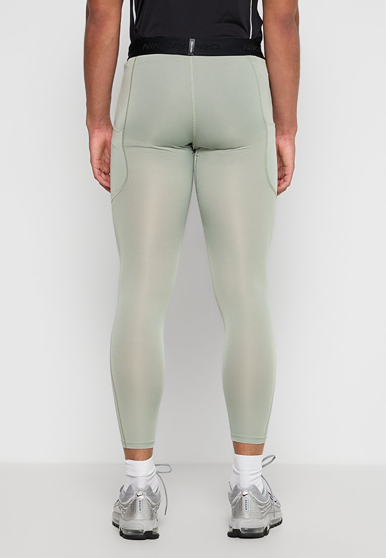 Hombre con leggings de compresión gris claro y zapatillas de correr plateadas, de pie con la espalda hacia el espectador frente a un fondo blanco liso.