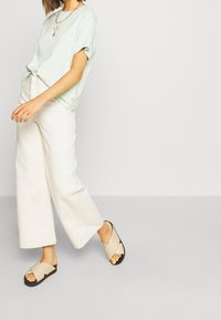 Svetle zelené tričko s krátkým rukávem, bílé kalhoty s širokými nohavicemi a béžové sandály na pásky s černou podrážkou. Minimalistický ležérní outfit.