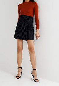 Ribbad röd tröja med långa ärmar, matchad med en marinblå rutig kjol med knytband i midjan och dubbelknappar som prydnad. Svarta strap-on klackar.