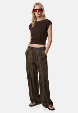 Vrouw met rood getinte zonnebril, een mouwloze zwarte crop top, losse geruite broek en zwarte teenslippers, staand met handen in de zakken.