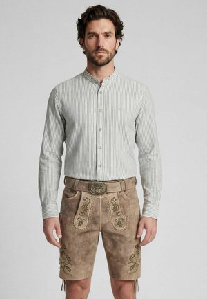 Mann trägt beige traditionelle Lederhosen mit Stickerei und ein grau-weiß gestreiftes Langarmhemd, steht vor einem einfarbigen Hintergrund.