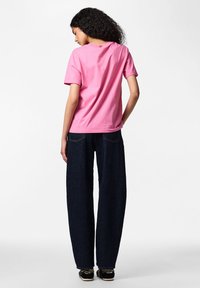 Femme aux cheveux bouclés portant un t-shirt rose à manches courtes, un jean foncé ample et des chaussures noires, debout dos à l'appareil photo.