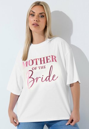 Blondynka ubrana w białą oversize koszulkę z różowym napisem „Mother of the Bride” i niebieskie dżinsy, stoi oparta o białą ścianę.