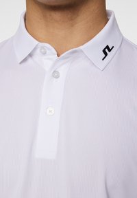 Weißes Poloshirt aus strukturiertem Stoff, mit zwei Knöpfen am Kragen und einem schwarzen Logo am linken Kragen.
