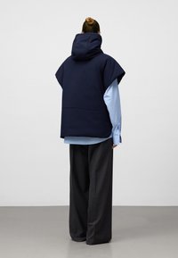 Granatowy bezrękawnik na kapturek nałożony na jasnoniebieską koszulę, z wysokim kołnierzem i luźnym krojem, zestawiony z szerokimi szarymi spodniami z teksturowanej tkaniny.