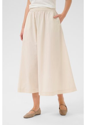 Kvinde iført beige vide bukser med elastisk talje og taupe flade sko, stående med den ene hånd i lommen.