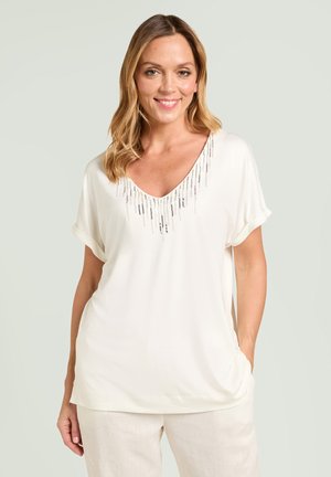 Femme souriante portant un haut blanc à manches courtes avec des perles argentées verticales près de l'encolure et un pantalon clair.