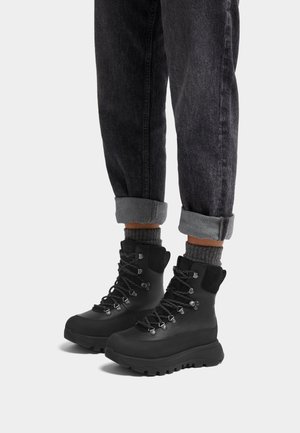 Schwarze wasserdichte Wanderstiefel mit einer robusten Sohle, Schnürdesign, gepolstertem Kragen und metallischen Verzierungen, kombiniert mit grauen hochgeschlagenen Hosen und Socken.