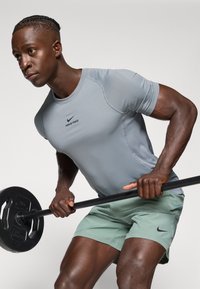 Camiseta deportiva ajustada de manga corta en gris con el logo "NIKE PRO", combinada con pantalones cortos de color verde claro; barra sostenida con un agarre texturizado marrón.