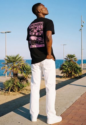 Mann steht nahe dem Strand mit Palmen, trägt ein schwarzes T-Shirt mit pinken Autografiken und weiße Hose, schaut nach oben gegen den klaren Himmel.