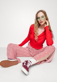 Top de botão vermelho canelado de mangas longas combinado com calças largas florais rosas. Modelo usa óculos de sol oversized verdes e ténis bordeaux.