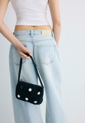 Oseba drži majhno črno kvačkano torbico z belimi okroglimi vzorci, oblečena v svetlo modre kavbojke in beli kratki top.