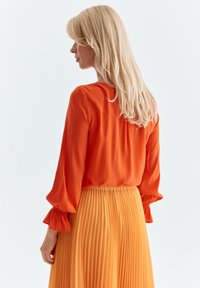 Orange Bluse mit Puffärmeln und gerafften Manschetten, kombiniert mit einem plissierten gelben Rock. Glatter Stoff mit einer leichten Textur.
