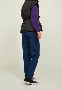 Černá prošívaná vesta přes fialový pletený svetr, v kombinaci s modrými džíny rovného střihu a černými vysokými teniskami. Jednoduchý, ležérní outfit.