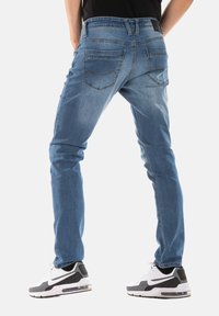 Jean en denim bleu avec une coupe slim, dotés de poches arrière et de coutures visibles. Associés à des baskets noires et blanches.