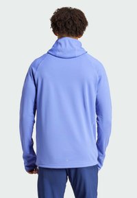 Mann mit lockigem Haar trägt einen hellvioletten Hoodie und dunkelblaue Hosen, steht mit dem Rücken zur Kamera vor einem einfarbigen, hellen Hintergrund.