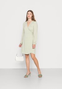 Vestido envolvente verde claro com mangas longas, tecido texturizado e detalhe de laço na cintura. Combinado com mules trançados verdes e uma bolsa branca.