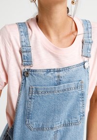 Ogrodniczki denimowe z regulowanymi metalowymi klamrami, z przednią kieszenią i jasnym zmyciem. W połączeniu z miękką różową koszulką.