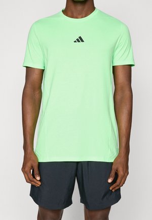 Man die een lichtgroen Adidas T-shirt met korte mouwen en zwarte korte broek draagt, staand tegen een effen achtergrond.