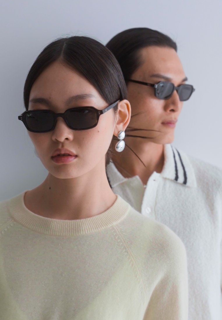 Lunettes de soleil noires rectangulaires portées par un modèle, associées à un pull jaune clair. Un autre modèle en arrière-plan porte une chemise blanche à col.