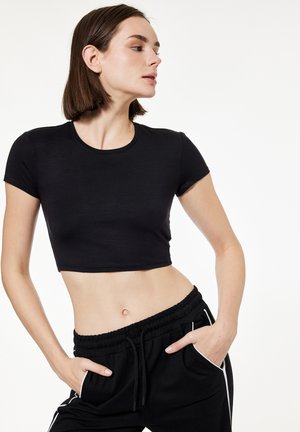 Ung kvinde iført en tætsiddende sort crop top og sorte bukser med snøring, med hænderne i lommerne, kigger til siden mod en hvid baggrund.