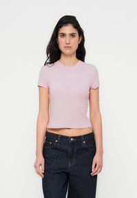 Vaaleanpunainen lyhyt t-paita, joka on valmistettu pehmeästä ja joustavasta kankaasta. Siinä on pyöreä pääntie ja lyhyet hihat. Yhdistetty tummansinisiin korkeavyötäröisiin farkkuihin.