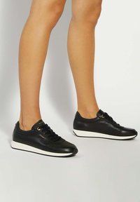 Legs wearing black leather sneakers with white soles, standing on a light gray surface against a plain background.