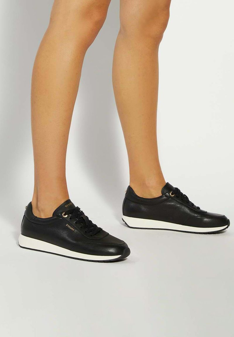 Legs wearing black leather sneakers with white soles, standing on a light gray surface against a plain background.