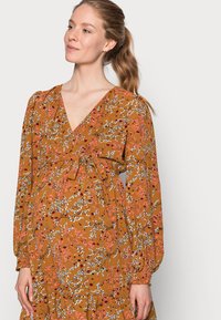 Vestido de manga longa, estilo envolvente, em amarelo mostarda, com padrões florais em rosa, branco e azul, com cintura franzida e bainha com volantes.