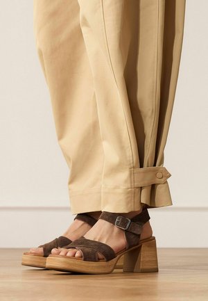 Voeten met bruine suède platformsandalen met houten blokhakken, gecombineerd met beige opgerolde broek tegen een neutrale achtergrond.