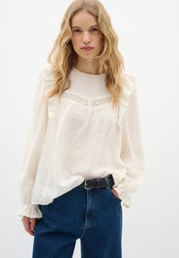 Camicia bianca a maniche lunghe con dettagli arricciati, inserto in pizzo al petto e vestibilità morbida, abbinata a jeans in denim blu a vita alta.