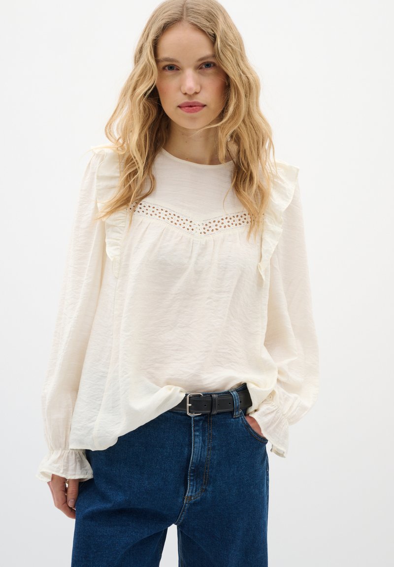 Camicia bianca a maniche lunghe con dettagli arricciati, inserto in pizzo al petto e vestibilità morbida, abbinata a jeans in denim blu a vita alta.