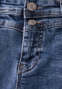 Tejido de denim azul oscuro con costuras visibles, cierre de botón de metal y detalle de bolsillo. Textura suave con ligero desvanecimiento.