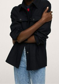 Personne portant une chemise noire boutonnée avec les manches retroussées et un jean bleu clair, bras croisés devant un fond uni.