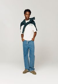 Hvit t-skjorte med korte ermer og grønn tekst, kombinert med blå jeans og brune sko. Mørkegrønn genser lagt over skuldrene.