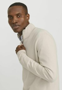 Šviesiai rudos zip-up megztinis su raukiniais ant apykaklės ir rankovių, puoštas tekstūruotu mezginiu ir sluoksniuotu juostuotu marškinėliais po juo.