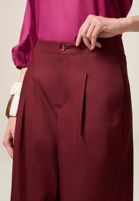 Bordeauxkleurige broeken met plooien, met een knoopsluiting op de tailleband. Een roze blouse met elastische manchetten is gedeeltelijk zichtbaar.