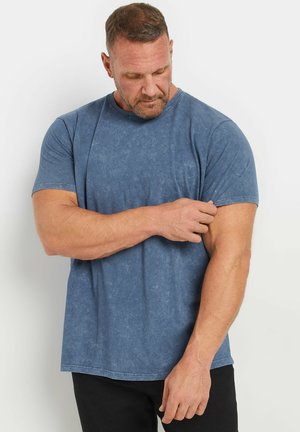 Man in blauw shirt met korte mouwen die zijn mouw afstelt, staand tegen een effen witte achtergrond.