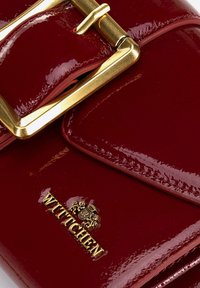 Červená lakovaná peněženka s zlatým přezkovým detailem a embosovaným logem. Hladká textura, kompaktní design a jasně definované okraje.