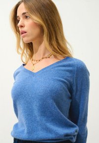 Blå V-ringad tröja i mjukt material med avslappnad passform. Har en subtil struktur och bärs med lager av guldhalsband.