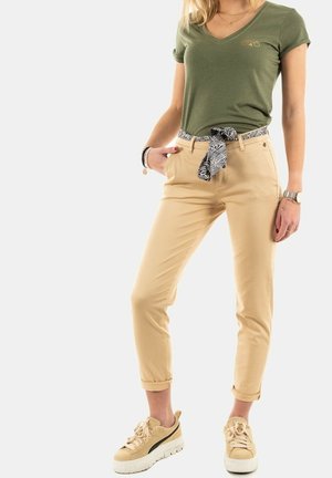 Kobieta w oliwkowozielonej koszulce z dekoltem w serek, beżowych spodniach podwiniętych do góry z wzorzystym paskiem, beżowych trampkach i bransoletkach na jednym nadgarstku.