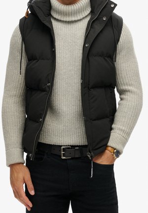 Homme portant un pull à col roulé côtelé gris clair, un gilet sans manches matelassé noir, une ceinture noire, un pantalon foncé et une montre-bracelet en or.