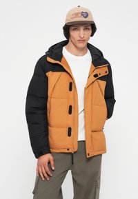 Mlađi muškarac nosi puffer jaknu u bež i crnoj boji, bijelu majicu, khaki cargo hlače i bež kapu s natpisom i logotipom.